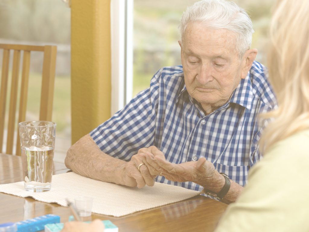 En la imagen, un señor mayor está sentado en la mesa de su casa, con un vaso de agua y varios pastilleros. Está organizando las pastillas en su mano, mientras una mujer que vemos en escorzo, posiblemente su hija, lo supervisa. Asegurar una correcta toma de los tratamientos, sobre todo en personas mayores polimedicadas, es una tarea que puede requerir ayuda de los familiares cuidadores.