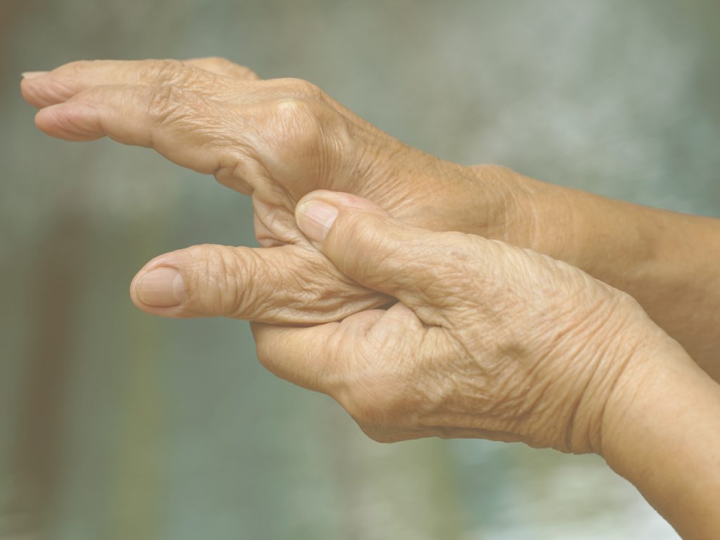 En la imagen, vemos las manos de una persona mayor. Se observa cierta deformidad en los dedos, posible consecuencia de la artrosis, y la persona se toca con una mano la base del pulgar de la otra, en lo que podría ser un gesto que traslada dolor o rigidez. Para aliviar el dolor de la rizartrosis y prevenir su avance, podemos realizar ejercicios sencillos con objetos cotidianos o seguir una sencilla tabla de ejercicios.