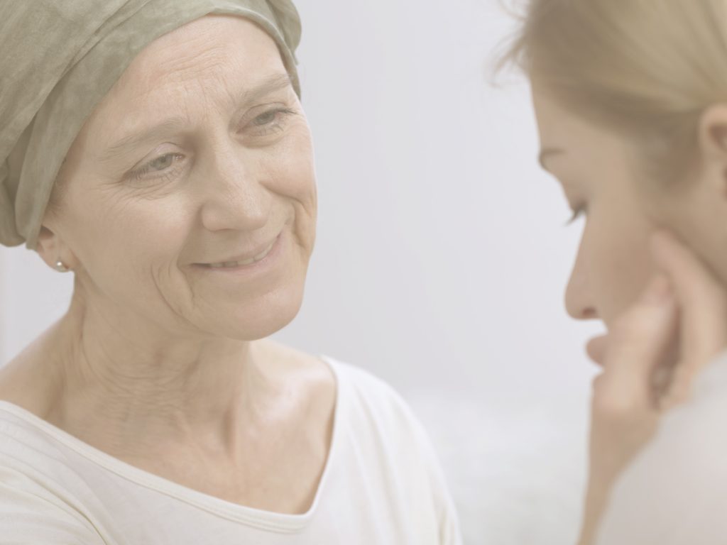 En la imagen vemos a una mujer con un pañuelo en la cabeza sonriendo mientras mira a otra mujer que se encuentra frente a ella y le acaricia el rostro con cariño. Cuando alguien está enfermo de cáncer, hablar sobre cómo nos sentimos y compartir nuestras preocupaciones sobre la enfermedad puede ayudar a aliviar la carga emocional y favorecer el acompañamiento en los momentos difíciles.