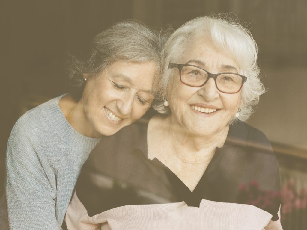 En la imagen vemos a dos mujeres, una mujer más joven que abraza con los ojos cerrados y con un gesto de cariño a la otra más anciana. La mujer mayor lleva unas gafas y sonríe contenta mientras recibe el abrazo de la otra mujer que parece ser de su familia.