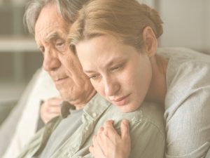 ¿Cómo gestionar el sentimiento de culpabilidad? Romper el mito del cuidador perfecto. En la imagen, vemos a una mujer que abraza a un hombre mayor, probablemente, su padre. Mientras él muestra un gesto ausente, ella tiene una expresión de profunda tristeza; parece que acabe de llorar.
