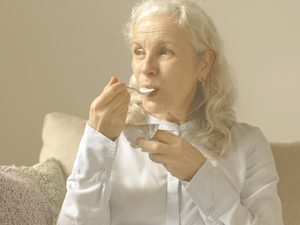 En la imagen vemos a una mujer mayor sentada en un sofá mientras come un yogur. ¿Cómo prevenir y ralentizar la osteoporosis en personas mayores? Una dieta rica en calcio, vitamina D y proteínas; la práctica de ejercicio regular; evitar hábitos como el tabaco y el alcohol y prevenir caídas son consejos clave en este sentido.