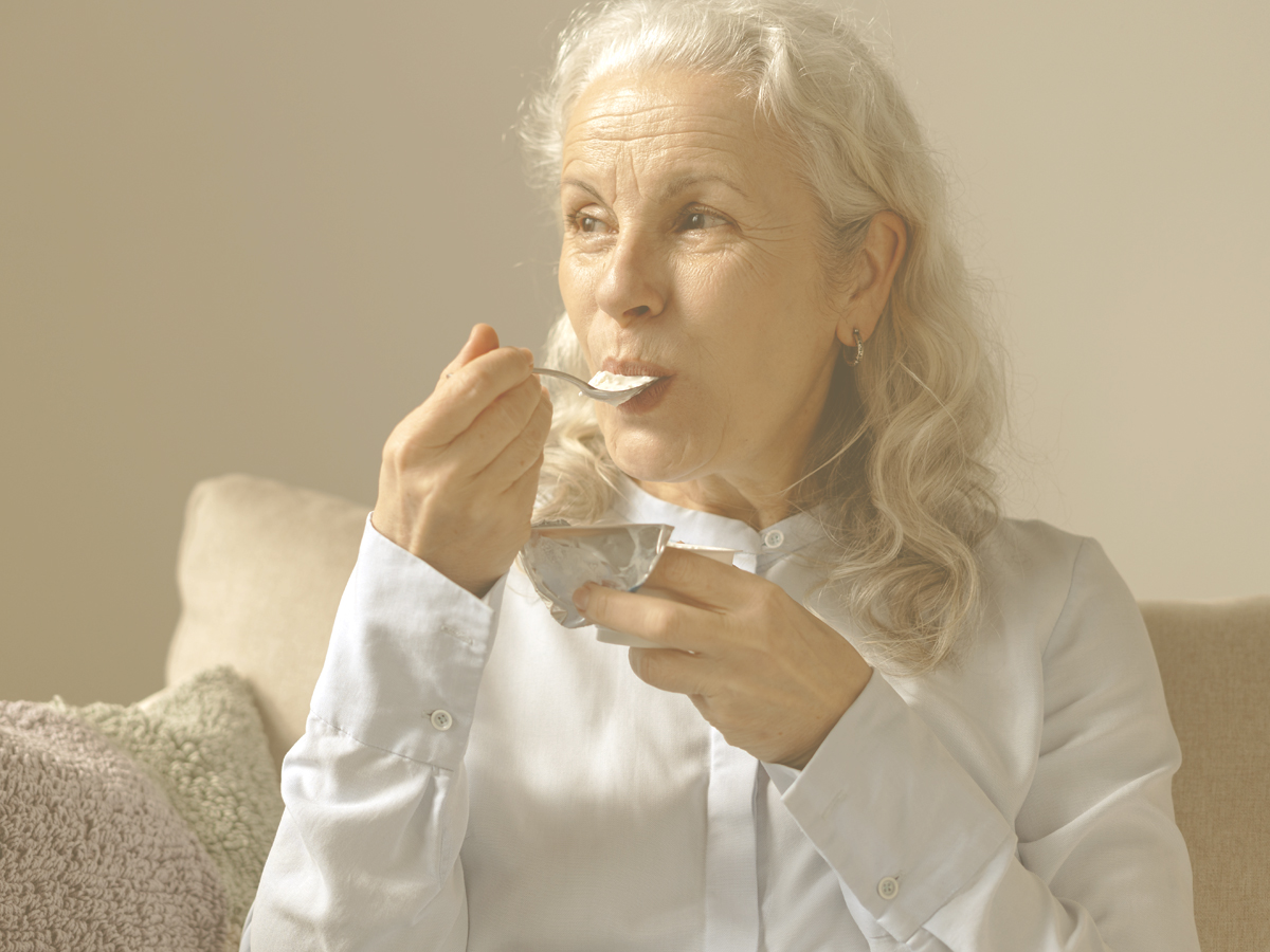 En la imagen vemos a una mujer mayor sentada en un sofá mientras come un yogur. ¿Cómo prevenir y ralentizar la osteoporosis en personas mayores? Una dieta rica en calcio, vitamina D y proteínas; la práctica de ejercicio regular; evitar hábitos como el tabaco y el alcohol y prevenir caídas son consejos clave en este sentido.