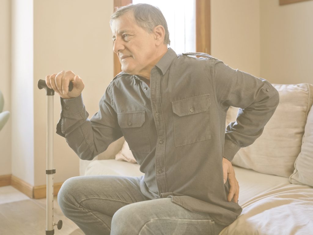 En la imagen vemos a un hombre mayor sentado en un sofá, apoyado en un bastón y llevándose una mano a la zona lumbar, como si sintiera dolor o molestias. La osteoporosis vuelve los huesos más porosos, debilitándolos e incrementando el riesgo de fracturas de fragilidad. El riesgo de desarrolarla puede reducirse con un estilo de vida saludable, pautas de ejercicio específicas y una dieta que garantice un correcto aporte de calcio y vitamina D.