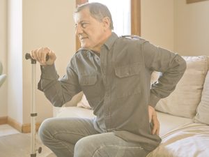En la imagen vemos a un hombre mayor sentado en un sofá, apoyado en un bastón y llevándose una mano a la zona lumbar, como si sintiera dolor o molestias. La osteoporosis vuelve los huesos más porosos, debilitándolos e incrementando el riesgo de fracturas de fragilidad. El riesgo de desarrolarla puede reducirse con un estilo de vida saludable, pautas de ejercicio específicas y una dieta que garantice un correcto aporte de calcio y vitamina D.