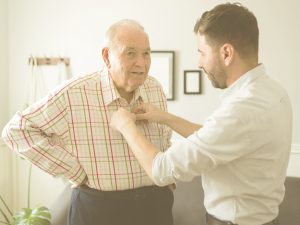 En la imagen, un señor mayor es ayudado por su hijo a atarse los botones de la camisa. Tiene un gesto tranquilo mientras ambos conversan. Seguir una serie de pasos hará que la tarea de vestir y desvestir a una persona en la cama o la silla de ruedas resulte más sencilla y cómoda.