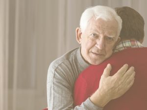 En la imagen, un anciano se abraza a quien podría ser su hijo, al que vemos de espaldas. El hombre mayor observa fíjamente la cámara, aunque su gesto es ausente. El cuidado de un familiar con alzheimer conlleva un gran impacto físico, psicológico y social. En este post, repasamos algunos de los aspectos a tener en cuenta a la hora de atender a una persona con esta demencia: cómo adaptar el hogar y las rutinas, estimularle y atender sus emociones, asumir sus cambios de humor y cuidarnos también nosotros en este proceso.