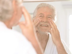 Claves para el cuidado de la piel en los mayores: la importancia de una correcta higiene e hidratación para evitar lesiones. En la imagen, vemos a un señor mayor que, vestido con un albornoz y sonriente, se observa en el espejo mientras se aplica una loción facial.
