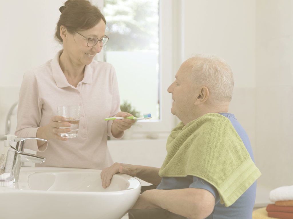 En la imagen vemos a un señor mayor en silla de ruedas frente al lavabo en su hogar. Una mujer de mediana edad, probablamente su hija, se dispone a cepillarle los dientes mientras ambos se sonríen. La higiene diaria de ojos, oídos y boca y dientes es importante para evitar problemas en personas mayores, sobre todo cuando estas no tienen autonomía o movilidad suficiente para llevar a cabo este cuidado por sí mismas.