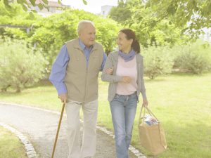En la imagen, vemos a un anciano con bastón que camina por un parque del brazo de una mujer más joven, probablemente, su hija. Ella lleva una bolsa de la compra y ambos conversan mientras ella le mira sonriente. Salir a pasear es una actividad que potencia la capacidad física en los mayores, así como ciertas habilidades cognitivas, y mejora su autoestima y socialización. Hay algunas pautas a tener en cuenta para hacer que esta actividad resulte saludable, y distintas ideas de rutas para hacerla más entretenida y estimulante.