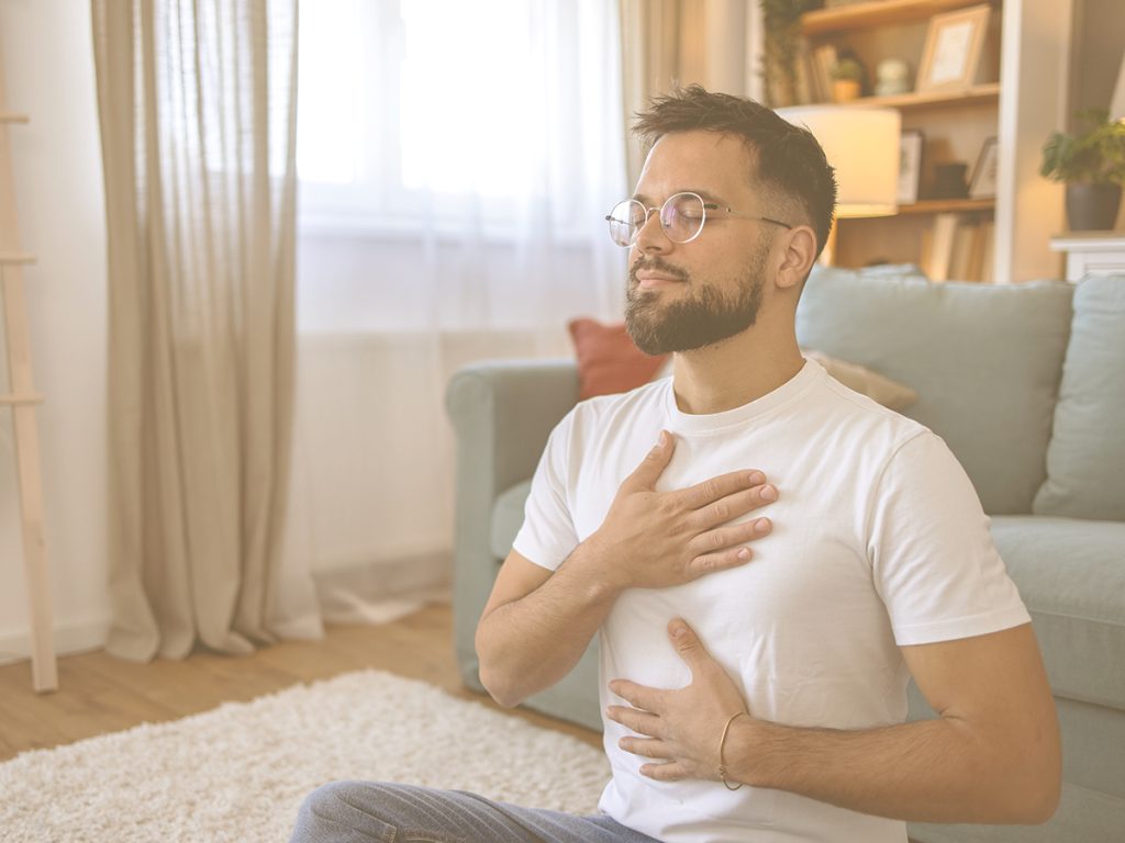En la imagen, un hombre joven se sienta en el suelo del salón de su casa, practicando ejercicios de relajación. Su gesto transmite calma y serenidad. La relajación puede ayudarnos a reducir el estrés y la ansiedad, mejorar el sueño, aliviar tensiones físicas y aumentar la concentración.