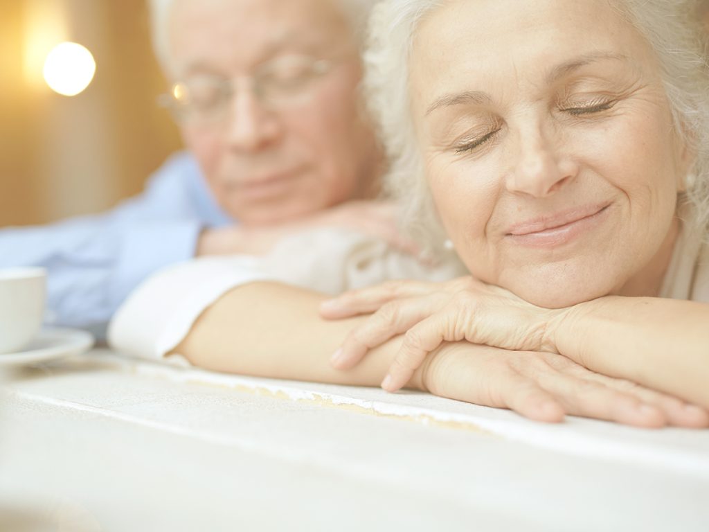 En la imagen, una mujer y un hombre mayor, apoyados sobre una mesa, sonríen con los ojos cerrados y un gesto de tranquilidad y serenidad. El mindfulness ayuda a conectar con uno mismo y a vivir el día a día de una manera serena y presente.