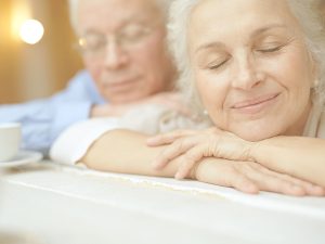En la imagen, una mujer y un hombre mayor, apoyados sobre una mesa, sonríen con los ojos cerrados y un gesto de tranquilidad y serenidad. El mindfulness ayuda a conectar con uno mismo y a vivir el día a día de una manera serena y presente.