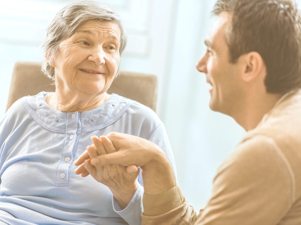 En la imagen, una anciana y un hombre más joven, probablemente su hijo, se dan la mano mientras se sonríen mutuamente. Cada etapa de cuidado lleva asociada retos, emociones y también diferentes posibilidades. Comprender cómo va evolucionado el cuidado puede ayudar a prepararse mejor, a organizarse con otros familiares o a pedir ayuda cuando se necesite.