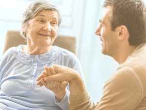 En la imagen, una anciana y un hombre más joven, probablemente su hijo, se dan la mano mientras se sonríen mutuamente. Cada etapa de cuidado lleva asociada retos, emociones y también diferentes posibilidades. Comprender cómo va evolucionado el cuidado puede ayudar a prepararse mejor, a organizarse con otros familiares o a pedir ayuda cuando se necesite.