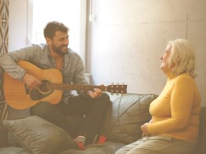 Musicoterapia significa “terapia a través de la música” y consiste en el uso profesional y guiado de la música con un objetivo claro: mejorar la salud y el bienestar de las personas. En la imagen, vemos a un hombre tocando la guitarra mientras una mujer mayor, tal vez su madre, le escucha y sonríe.