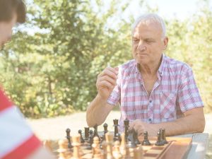 Juegos clásicos para jugar con personas mayores en casa, fomentar la interacción social y disfrutar de momentos de ocio juntos. En la imagen podemos ver a un hombre de unos 70 años jugando una partida de ajedrez con otra persona en un parque soleado.