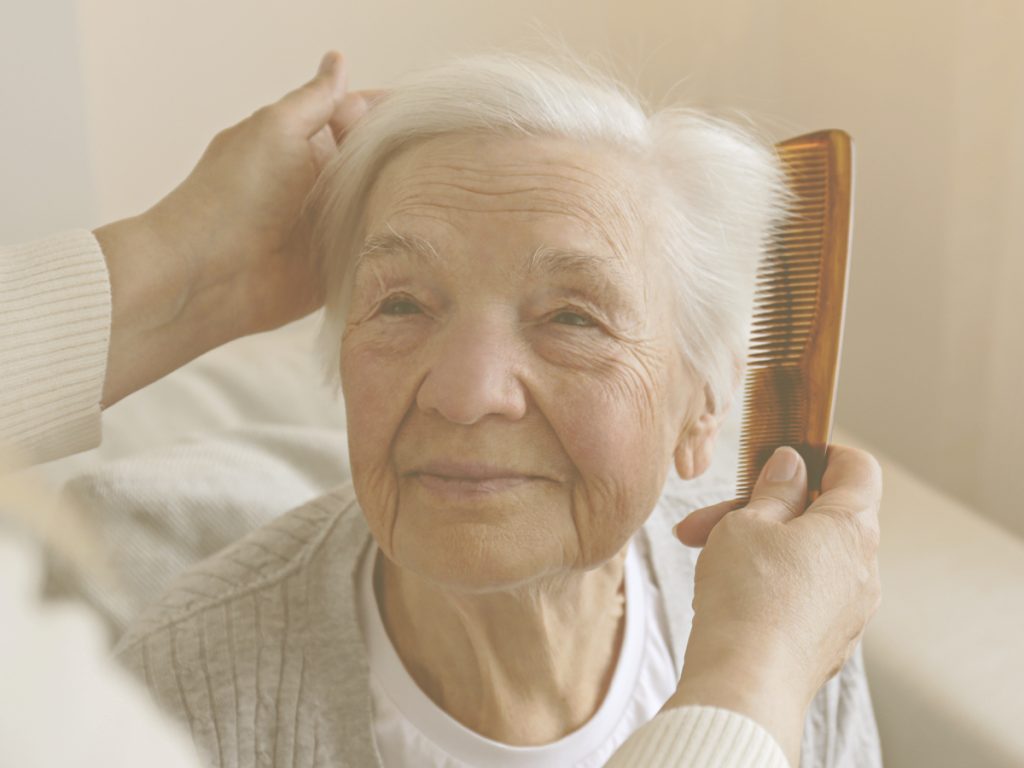 En la imagen vemos un primer plano de una mujer mayor de pelo corto que está sentada en la cama. Unas manos le están peinando con delicadeza. La rutina de higiene en personas que permenecen largos periodos de tiempo en la cama, debe incluir el lavado del cabello. Esto contribuye al bienestar y comodidad de nuestro familiar, reforzará su autoestima y sus relaciones familiares y sociales.