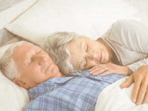 Sueño, insomnio y descanso en personas mayores: consecuencias del insomnio y recomendaciones para mejorar el sueño nocturno en ancianos. En la imagen podemos ver una pareja de personas mayores durmiendo en su cama. La mujer está recostada sobre el pecho del hombre.